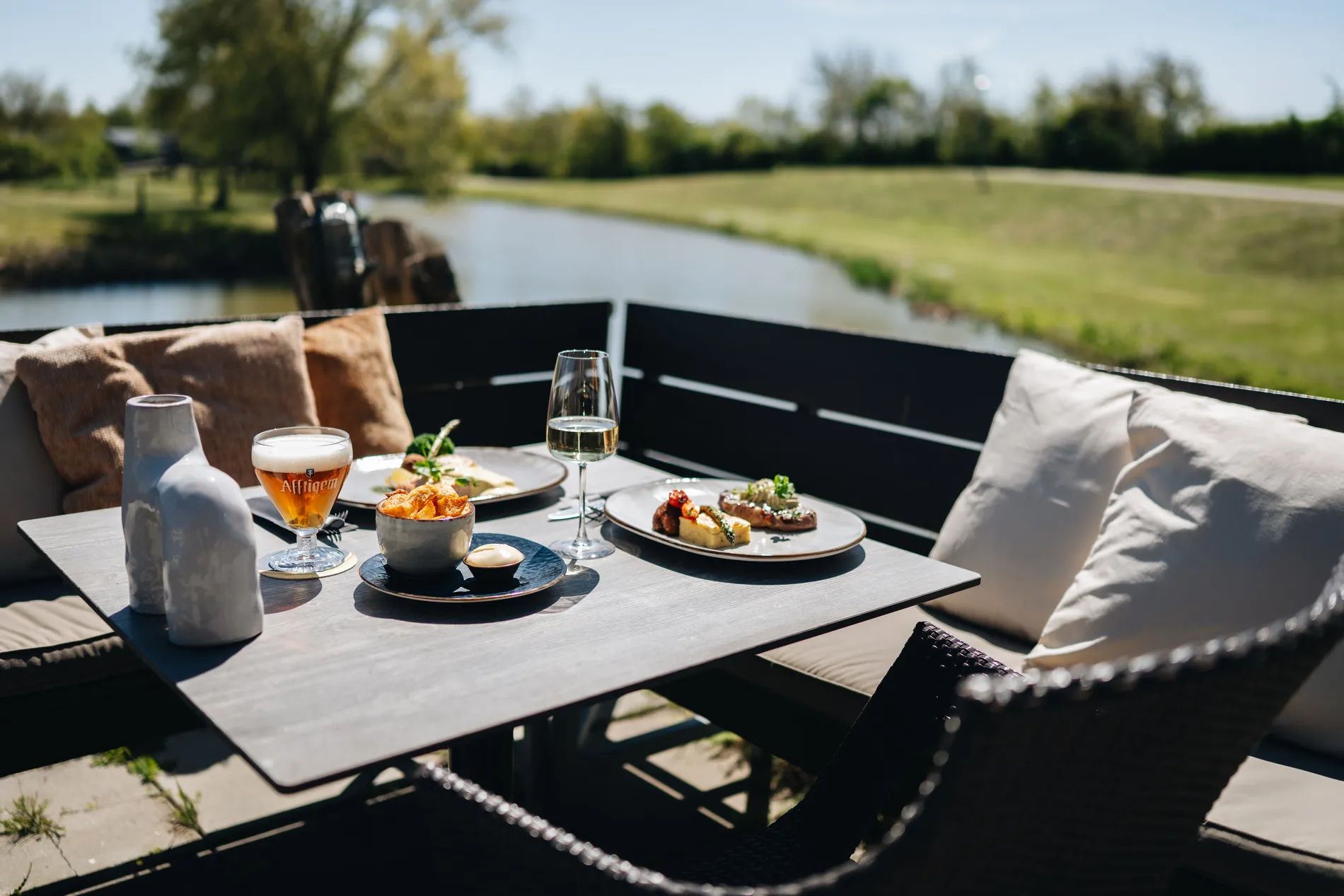 Een gedekte loungetafel op het buitenterras aan het water met een glas Affligem bier, een glas witte wijn, een schaaltje frites en twee smaakvolle lunchgerechten.